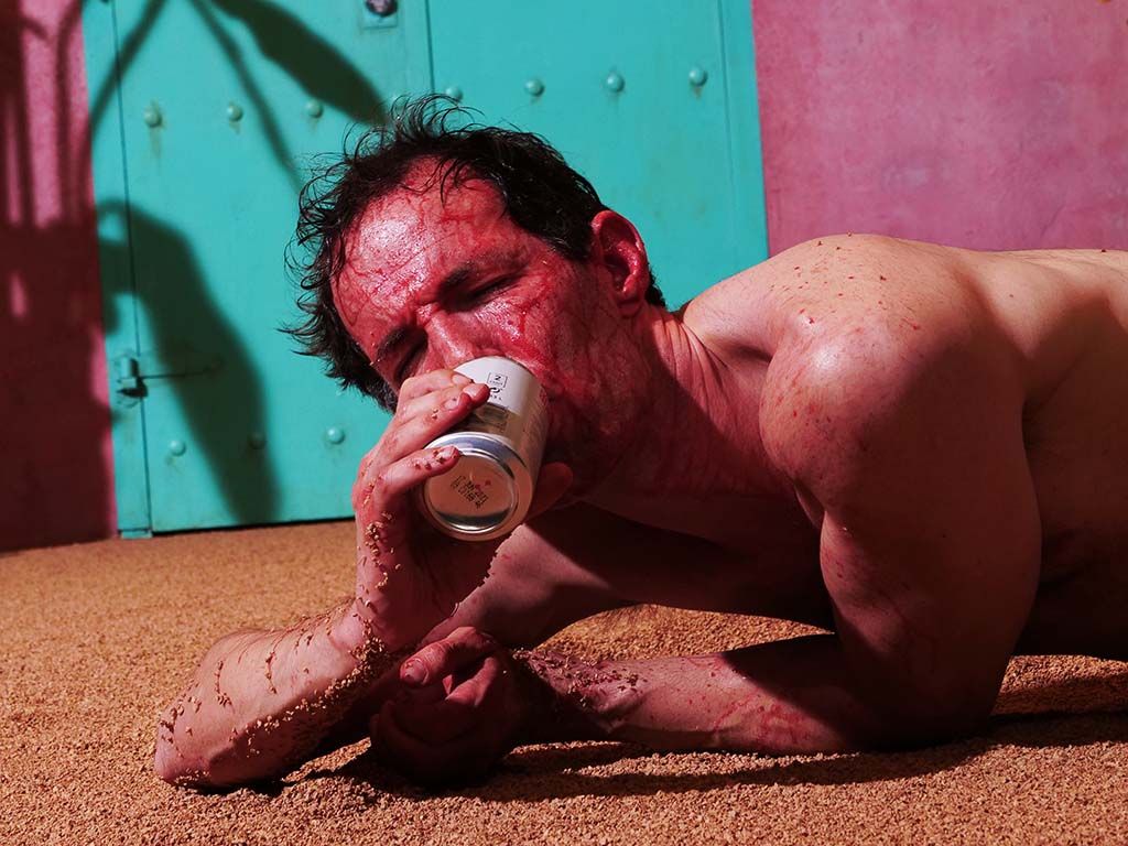 Bilde av mannlig skuespiller som ligger blodig på bakken og drikker noe fra en boks, fra forestillingen Licht und Liebe til Susie Wang