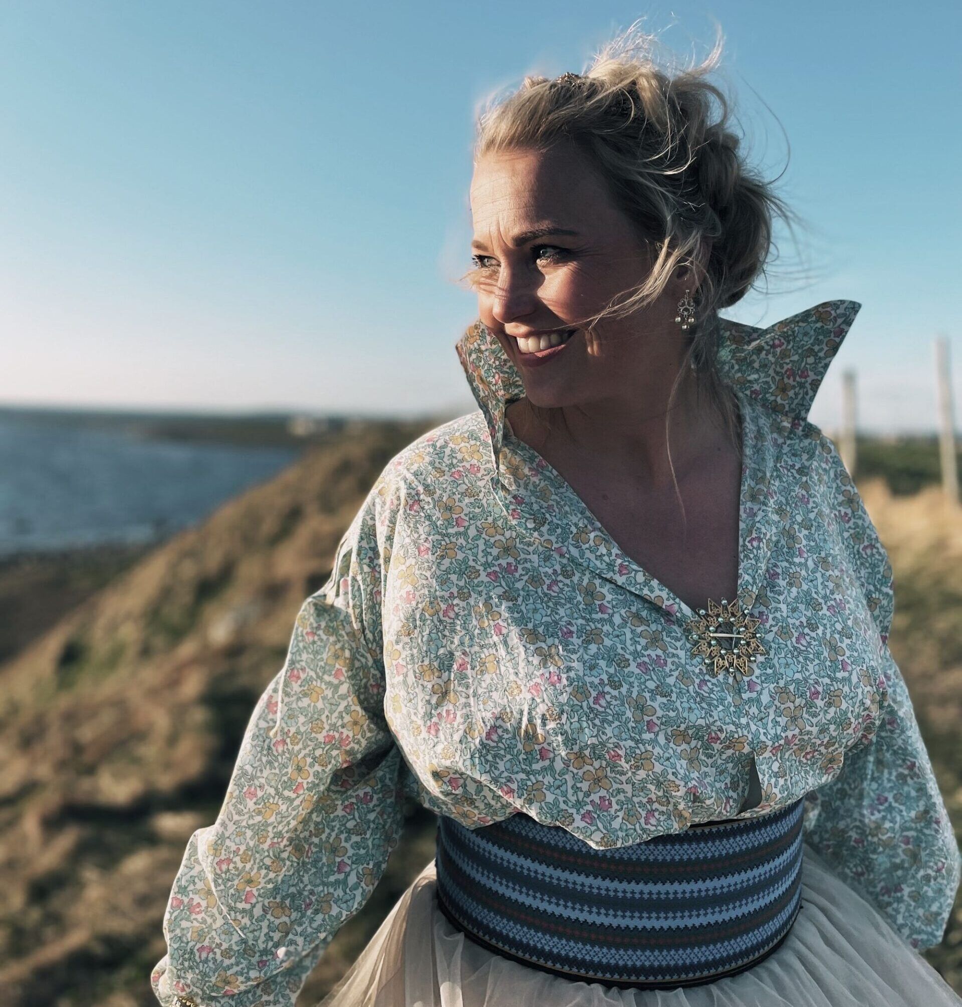 blid dame som står foran havet og smiler med solen i ansiktet