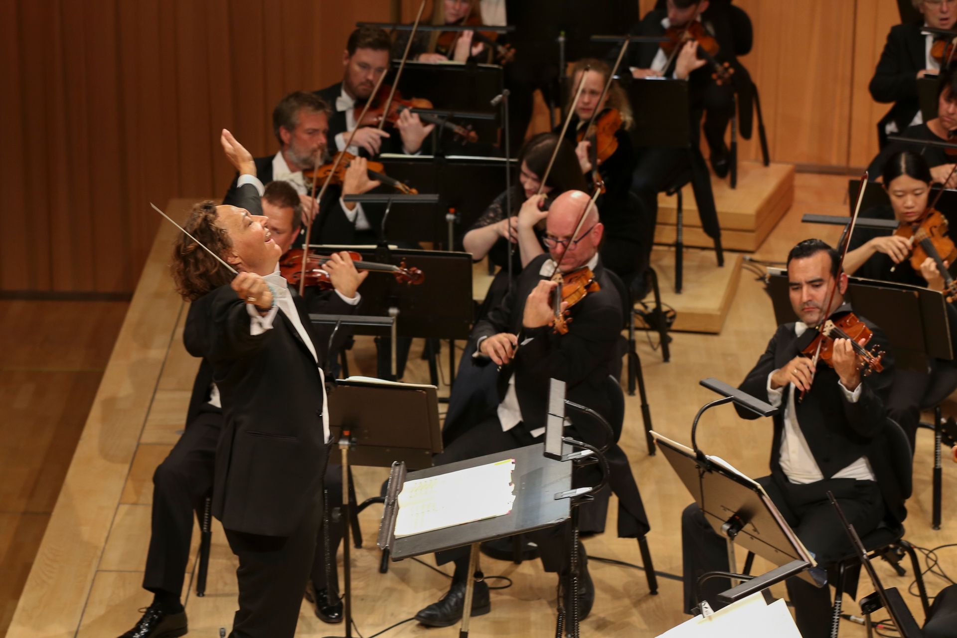 En kvinnelig dirigent som strekker armene i været. I bakrunnen spiller et symfoniorkester.