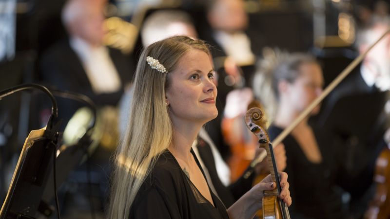 Kvinne med langt lyst hår som sitter i et orkester. Hun holder en fiolin.