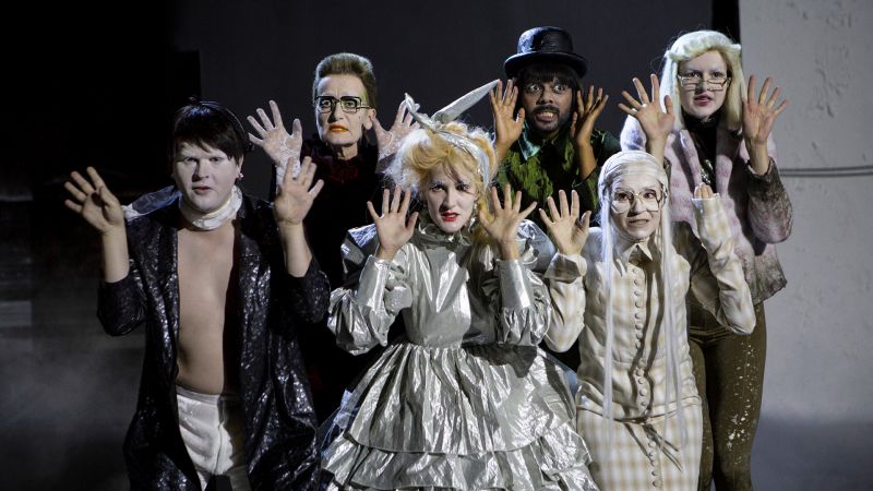 Bilde av seks personer i kostymer som holder hendene frem for seg og smiler anstrengt.