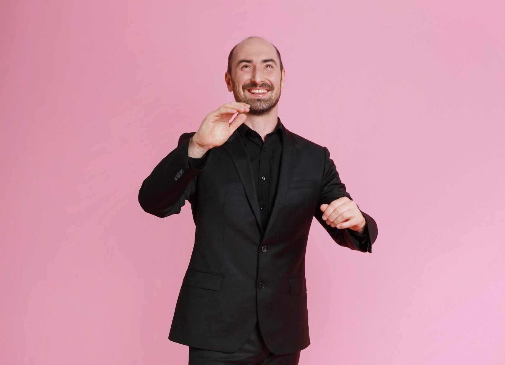 Blid mann som står i sort dress foran en rosa vegg og smiler fordi han elsker jobben sin