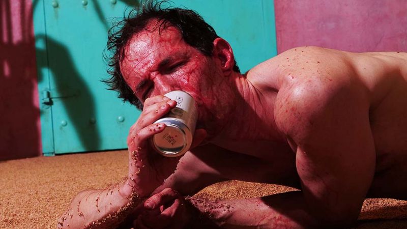 Bilde av mannlig skuespiller som ligger blodig på bakken og drikker noe fra en boks, fra forestillingen Licht und Liebe til Susie Wang