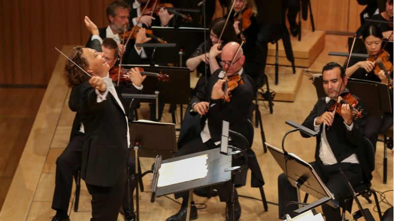 Bilde av kvinnelig dirigent som smiler mens hun strekker armene ut. Musikere som spiller i bakgrunnen.