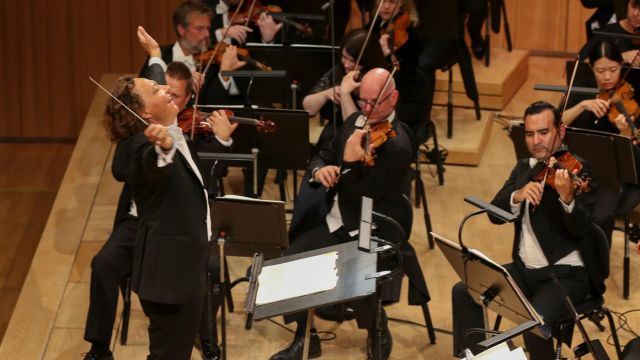 Bilde av kvinnelig dirigent som smiler mens hun strekker armene ut. Musikere som spiller i bakgrunnen.