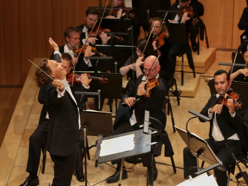 Kvinnelig dirigent som smiler mens hun dirigerer. I bakgrunnen ser du strykerne i orkesteret.