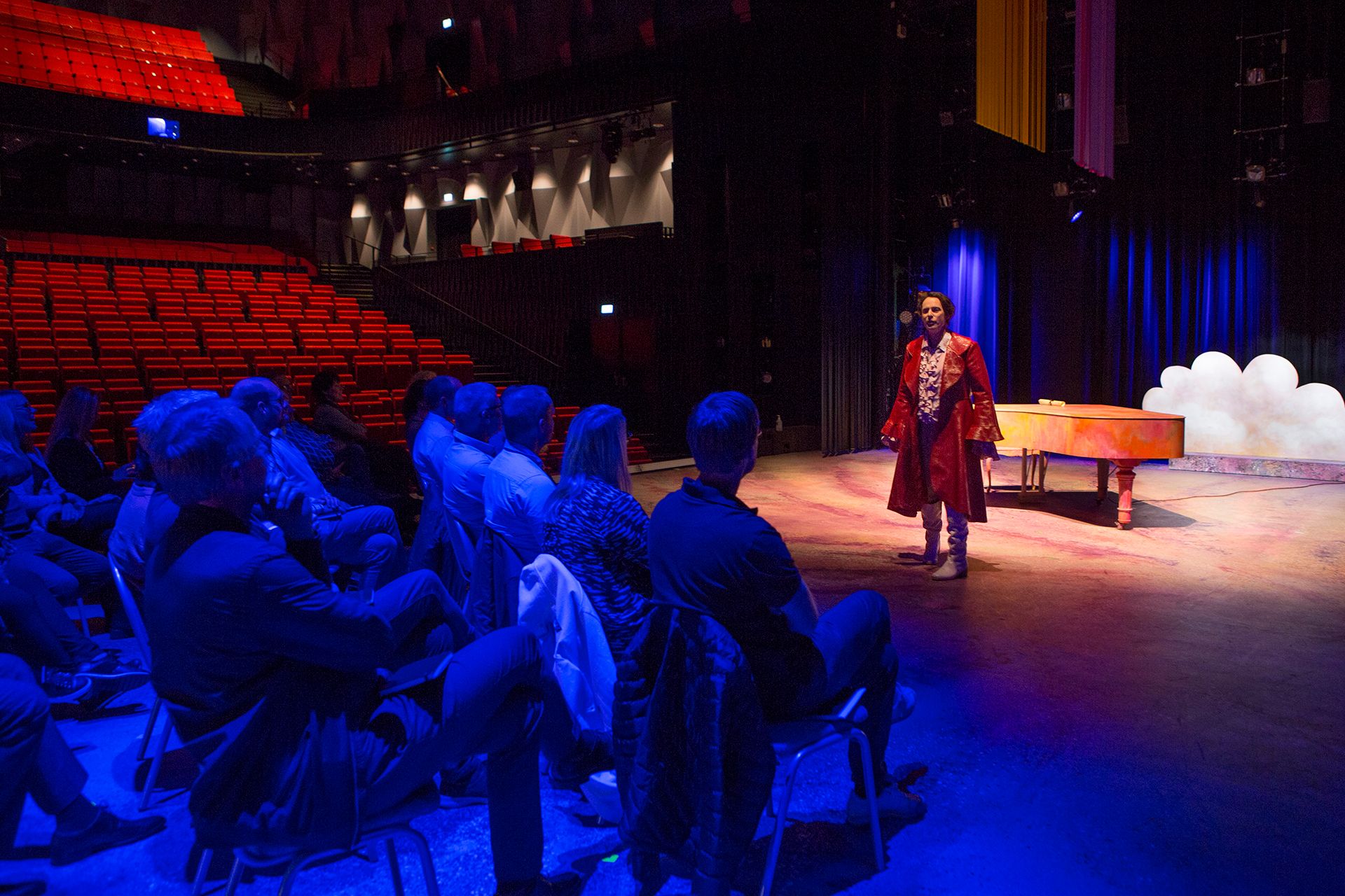 En gruppe mennesker som sitter på stoler på en teaterscenen. En mann i rød frakk står og prater til forsamlingen.