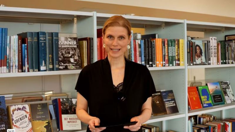 Bilde av Hilde Elise Bie, arrangementsansvarlig ved Grimstad bibliotek, foran en bokhylle ifbm en digital førsnakk for Idioten.