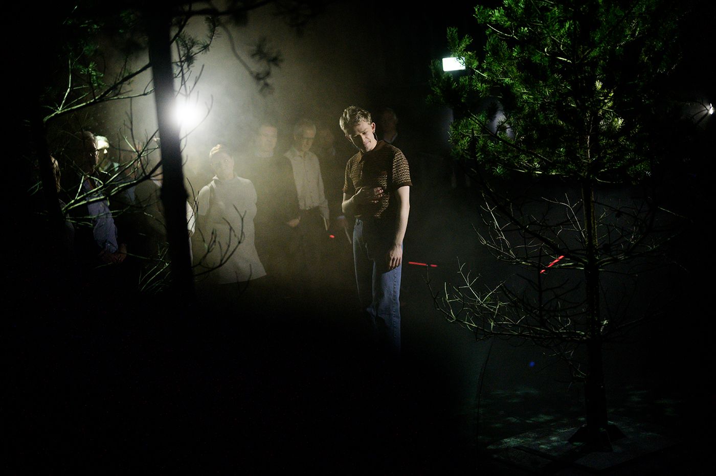 Bilde av en skuespiller som står mellom to furutrær. Publikum skimtes i bakgrunnen.