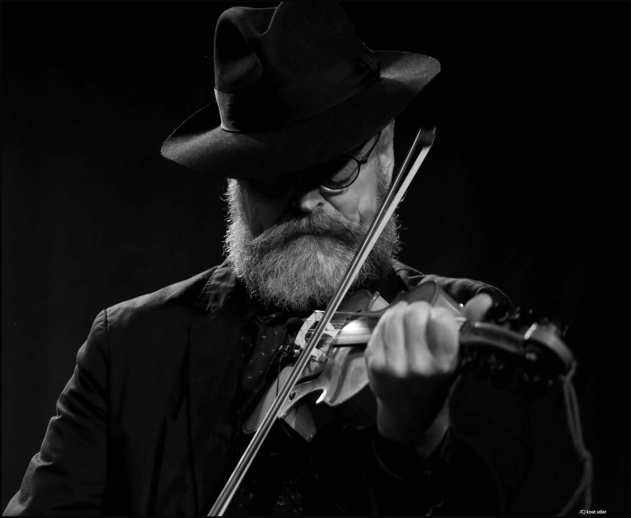 sorthvitt bilde av en mann med hatt og skjegg som spiller instrument foran sort bakgrunn