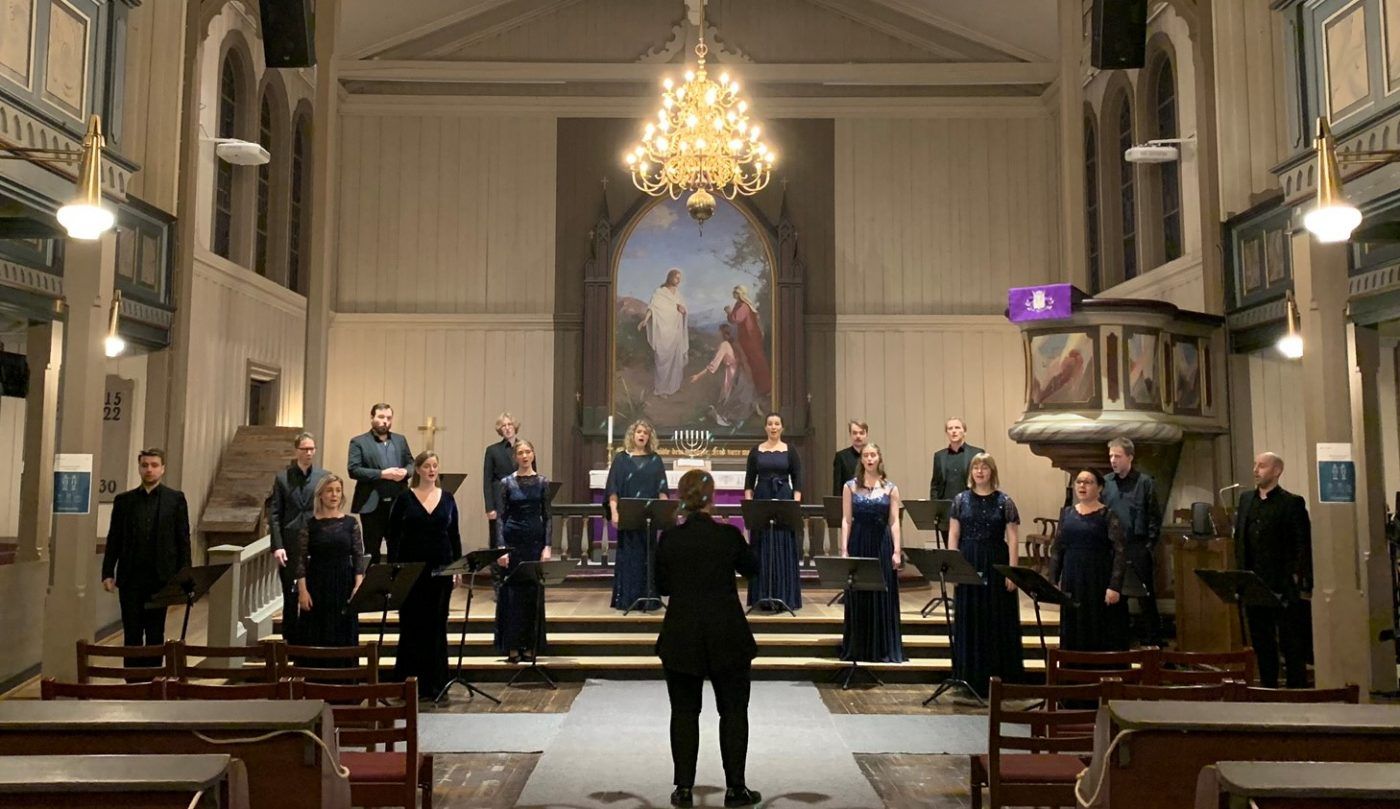 Bilde av sangere som er kladd i sort og som synger i en kirke. Kvinnelig dirigent står med ryggen til og dirigerer koret.