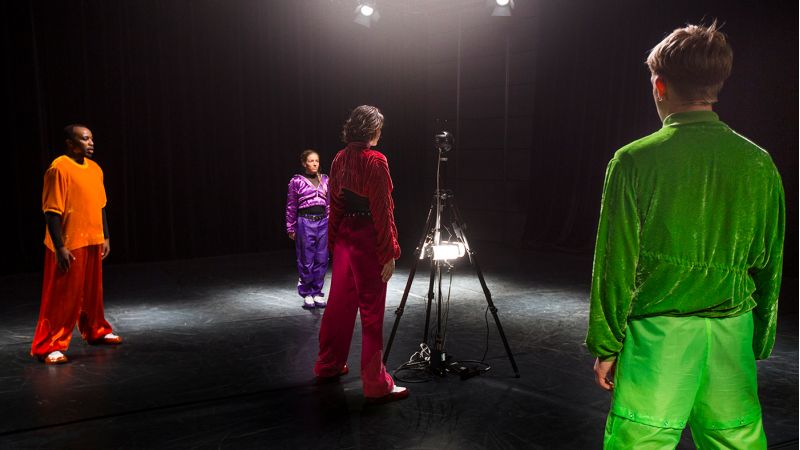 Bilde av fire unge voksne i fargerike treningsklær som står rundt et kamera.