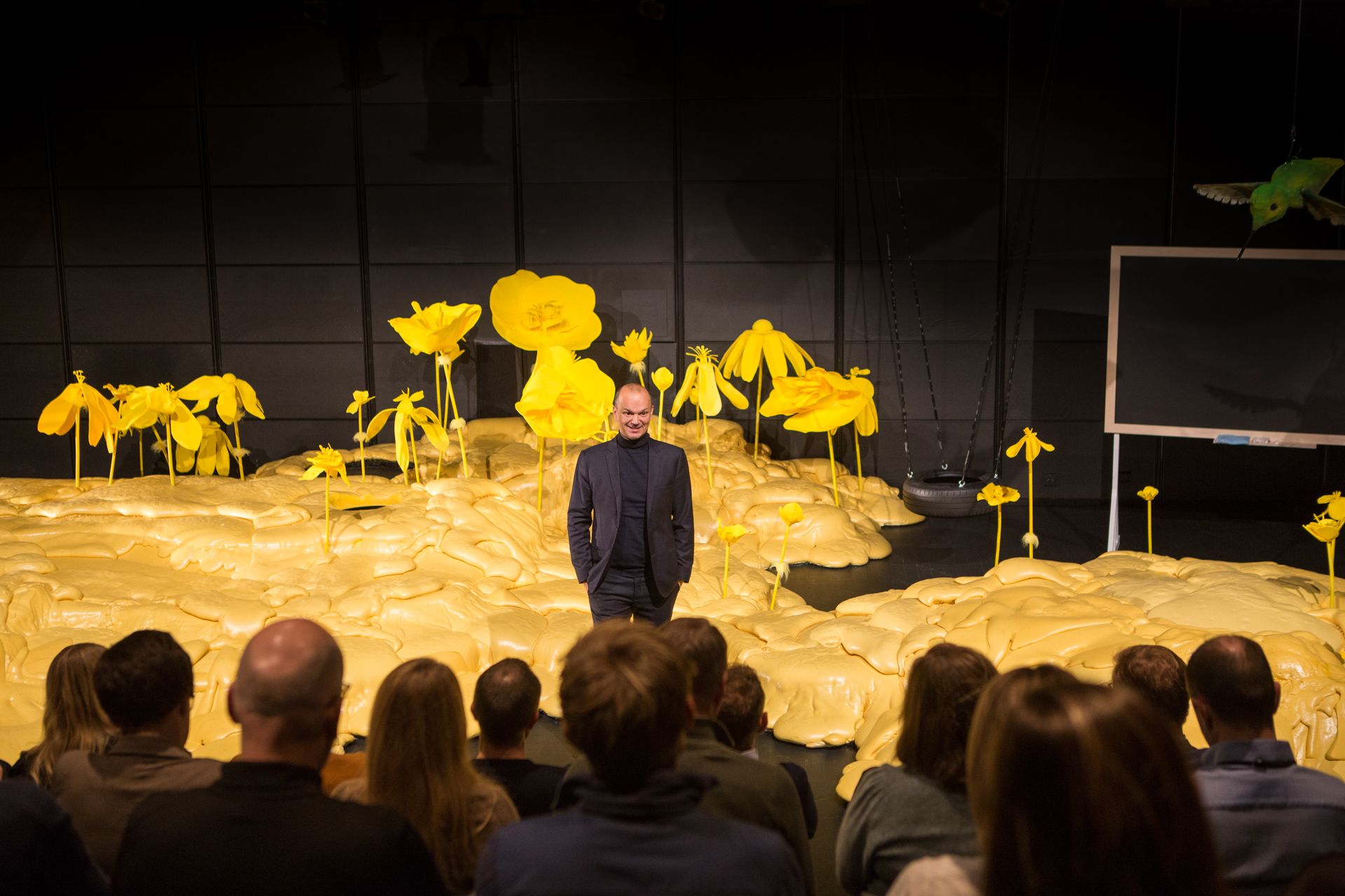 Mann i mørk dress som står på en scene og snakker til en forsamling. På scenen er det gult gulv og gule blomstre.