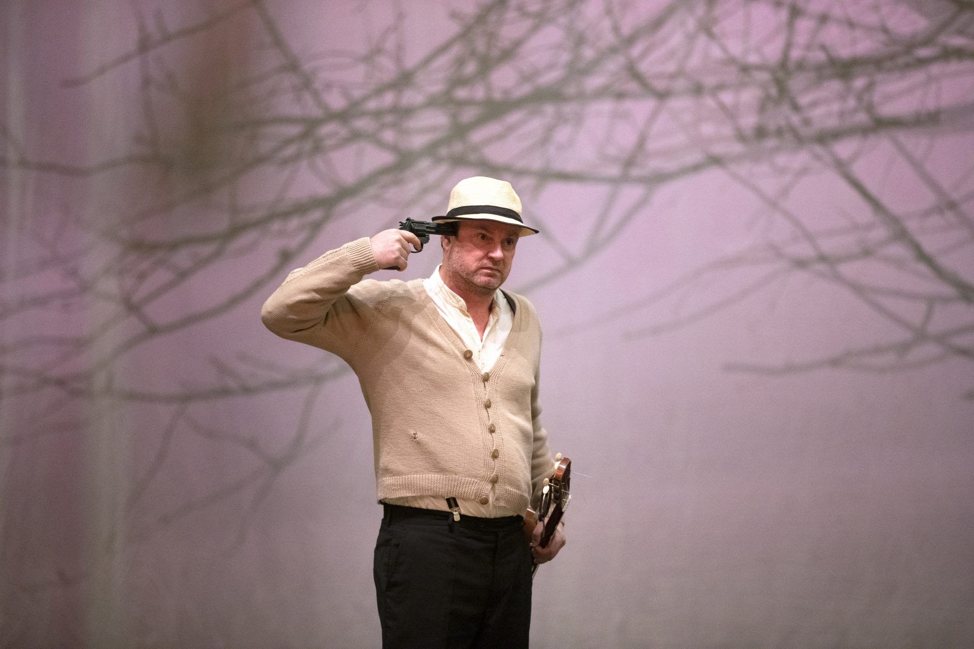 En mann med beige strikkejakke og lys hatt, holder en pistol mot hans hode. I bakgrunnen ser man greiner fra et kirsebærtre.