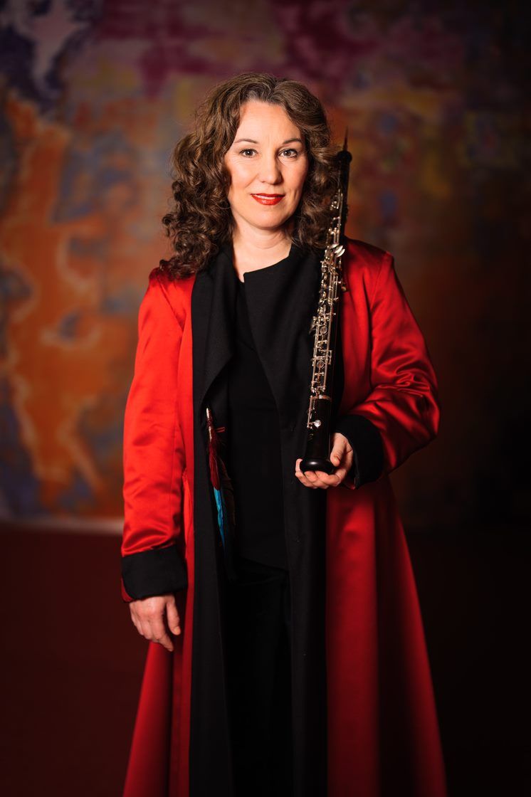 kvinne i rød og sort drakt som står oppstilt foran en fargerik vegg og smiler i kameraet mens hun holder sitt instrument i den ene hånden, bildet er varmt betont