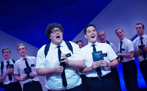 To unge menn i hvite kortermede skjorter og slips som synger. i hendene holder de Mormons bok. I bakgrunnen skimtes flere unge menn i samme bekldning.