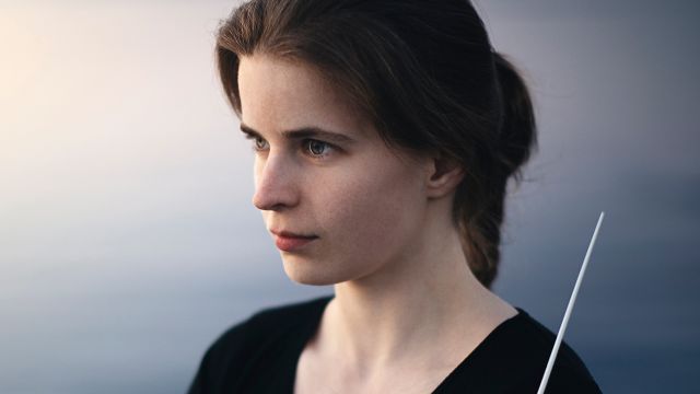 Bilde av kvinnne med langt mørkt hår i hestehale. Hun står ved sjøen og holder en dirigentstokk.