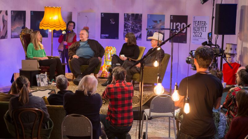4 personer som sitter og prater på en liten scene mens en gruppe mennesker sitter og ser på. Kameraoperatører er synlig i bildet.