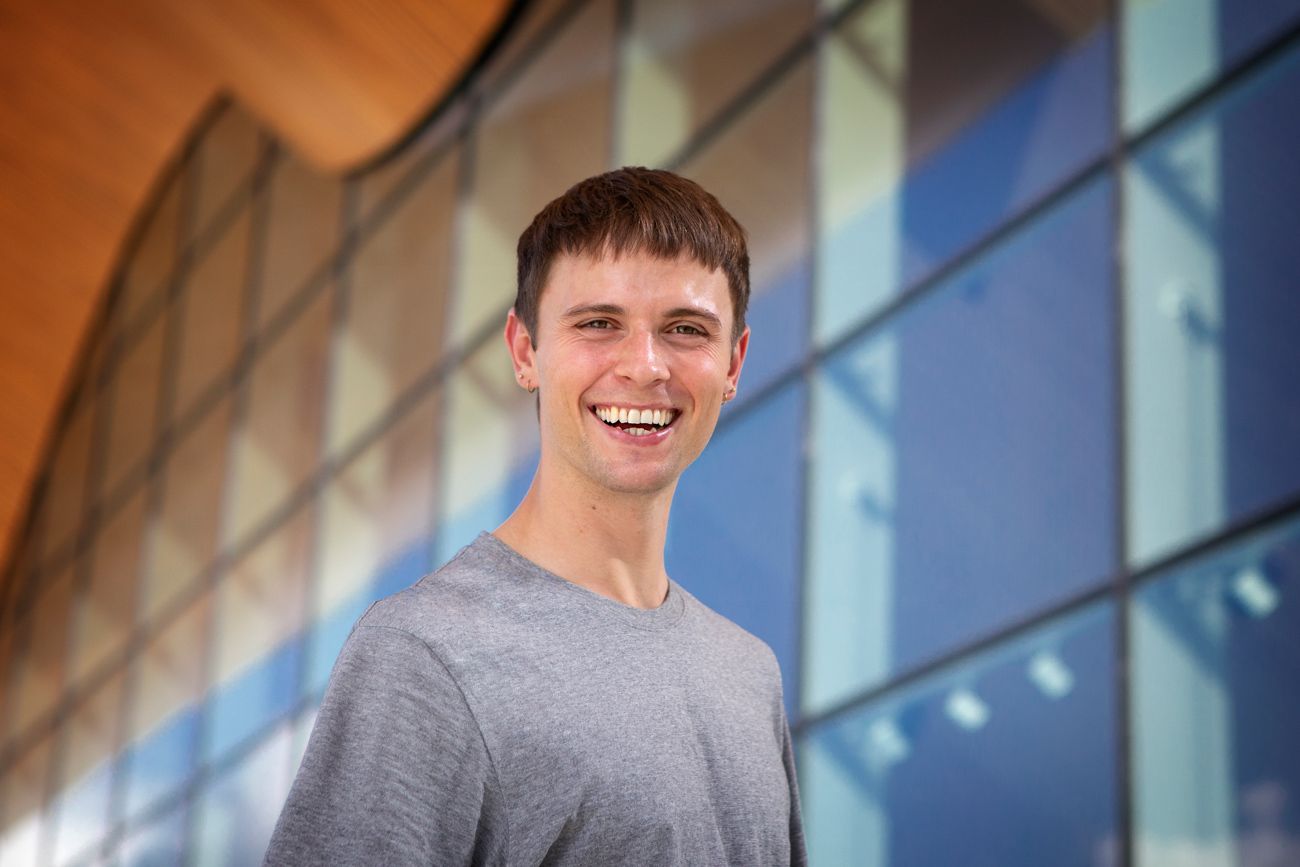 Bilde av ung mann som smiler foran et bygg med glassfasade.