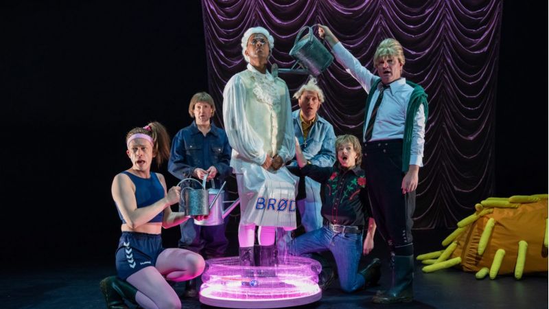 En mann i hvite klær og parykk står i et kar med vann. Rundt ham står fem personer som vanner ham med små vannekanner