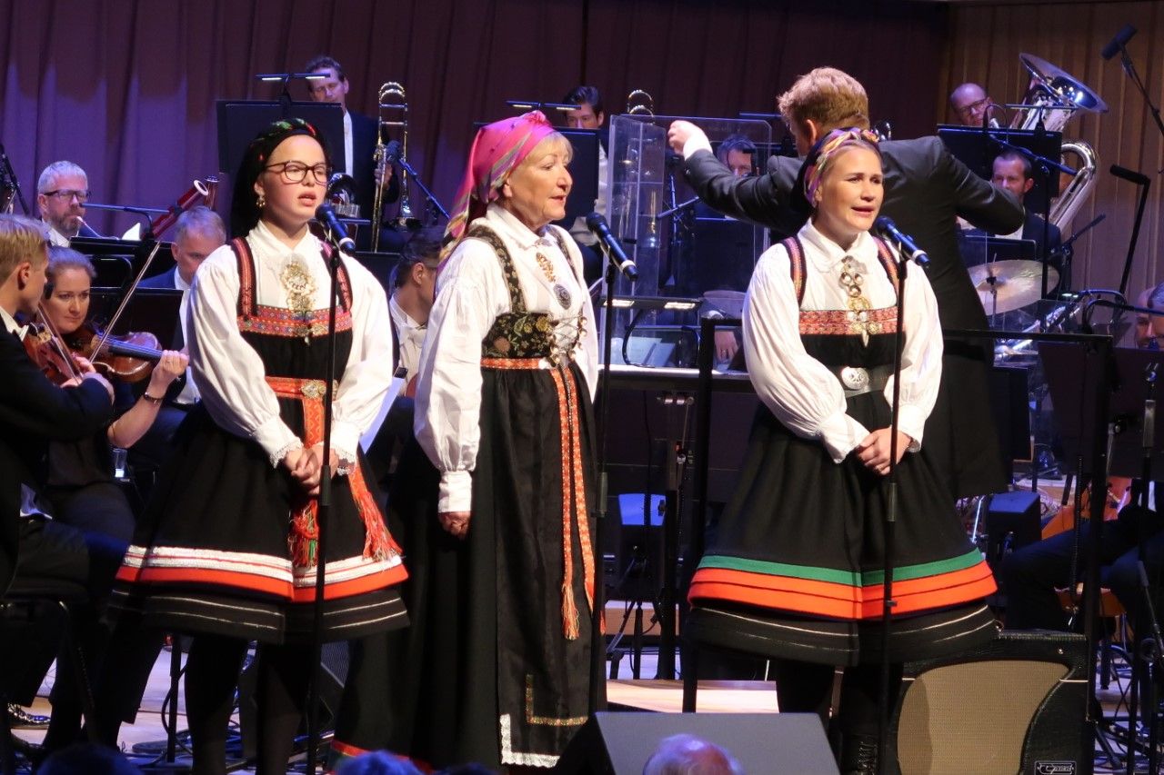 tre kvinner i nasjonaldrakt som synger på en scene med orkester