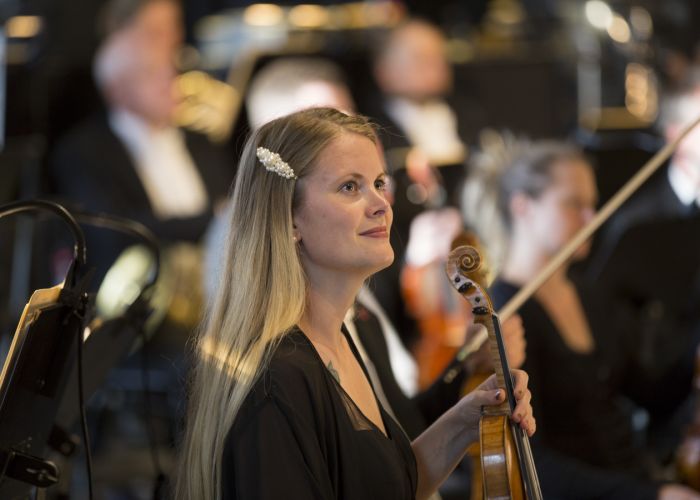 Kvinne med langt lyst hår som sitter i et orkester. Hun holder en fiolin.