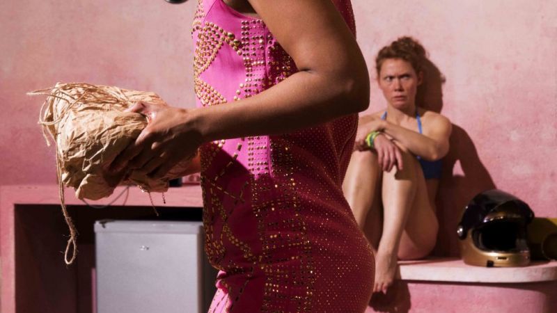 I forgrunnen ser du et menneske gå forbi i rosa kjole med en pakke i hendene. I bakgrunnen en kvinne i bikini som sitter på en benk med en scooter-hjelm ved siden av seg.