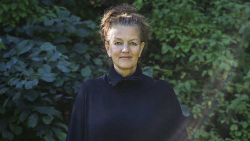 Bilde av kvinne med sort genser og mørkt hår. Stående utendørs fremfor trær med grønne blader.