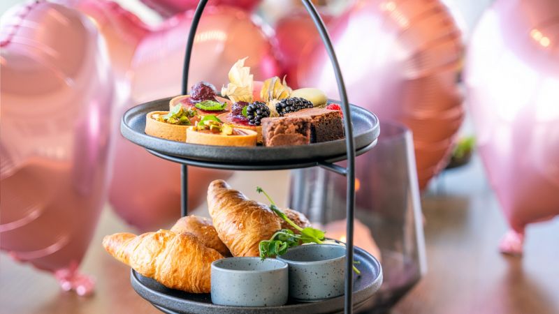 bilde av tre etasjers fat med ulike retter og dessert. Hjerte-heliumballonger i bakgrunnen. Bildet har preg av valentines dagen.