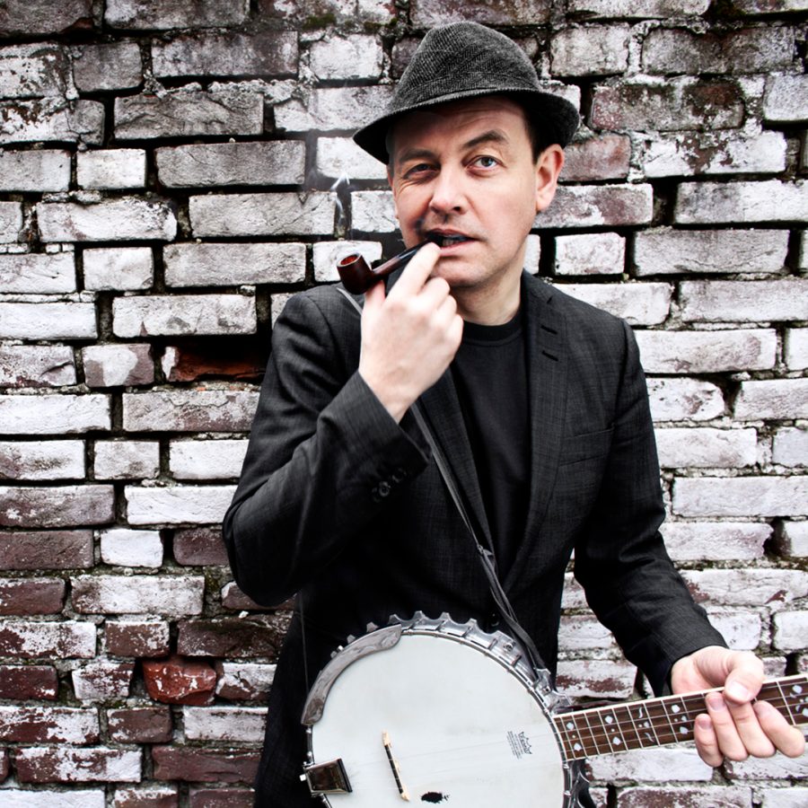 Bilde av mann med hatt og pipe som holder en banjo. I bakgrunnen er det en mursteinsvegg.