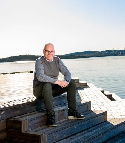 Mann med briller som sitter på en trapp som går ned til vannet. I bakgrunnen er det sjø og skjærgård.