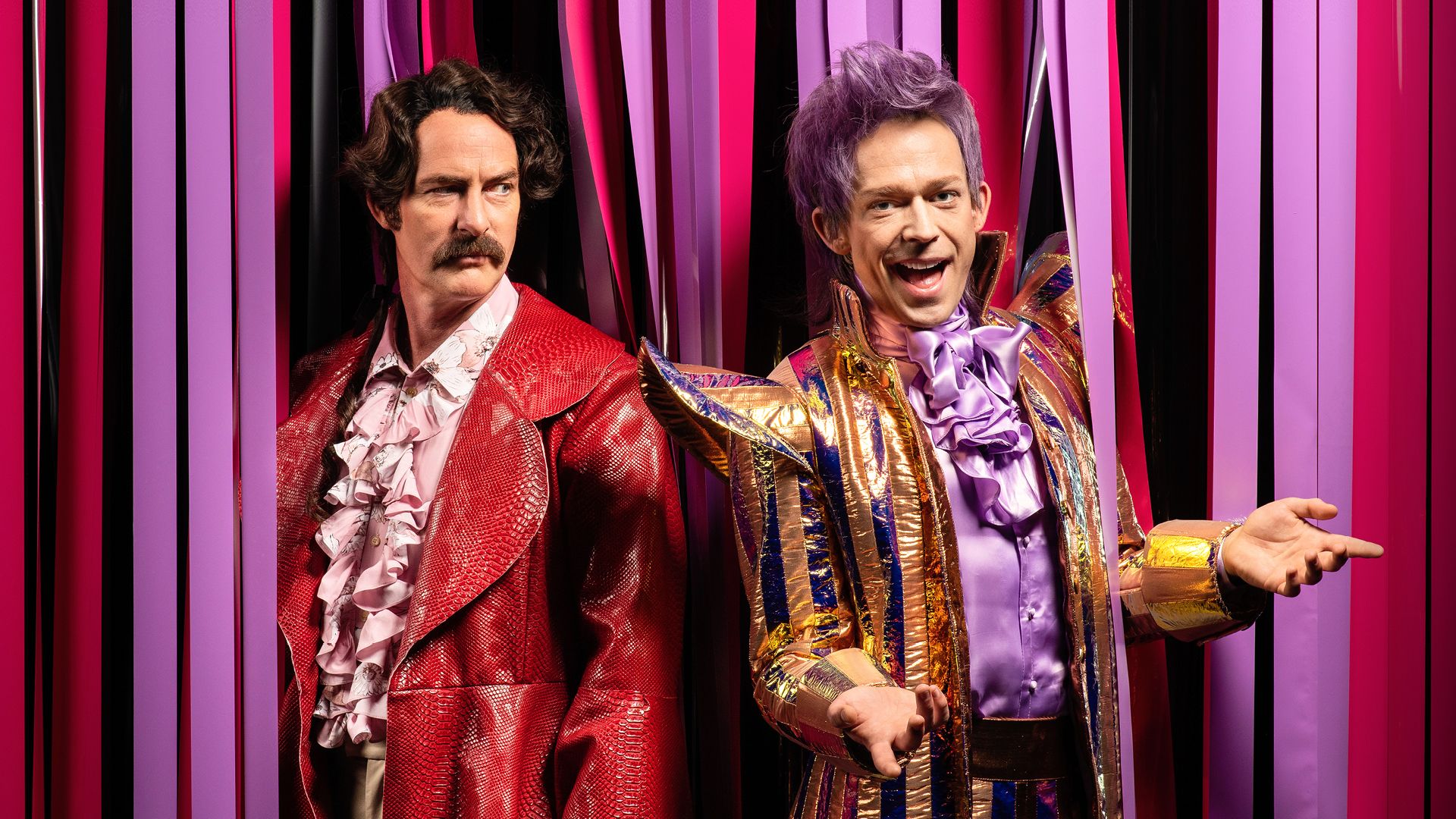 To medd i fargerike klær som ligger på bakken blant sorte remser. Bildet er tatt ovenfra og de ser rett inn i kameraet.