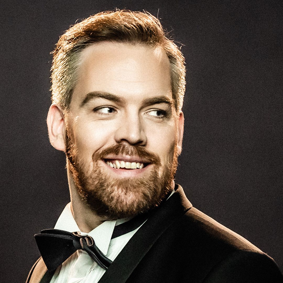 image of a man with short blond hair and beard smiling as he looks over his shoulder. He is wearing a white shirt, black bow tie and black jacket.