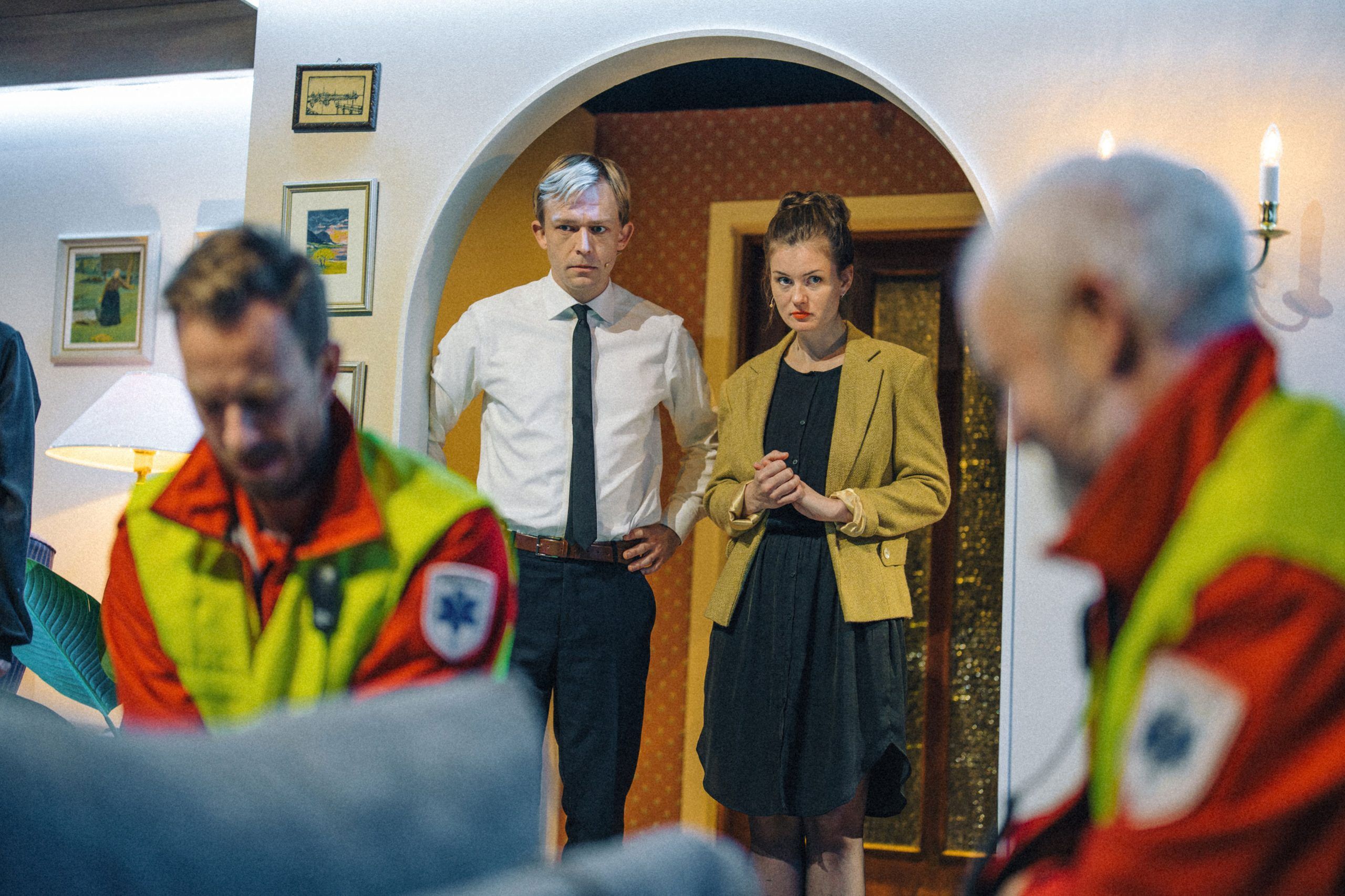 Espen og Elisabeth står nervøst i døråpningen og ser på to ambulansearbeidere.
