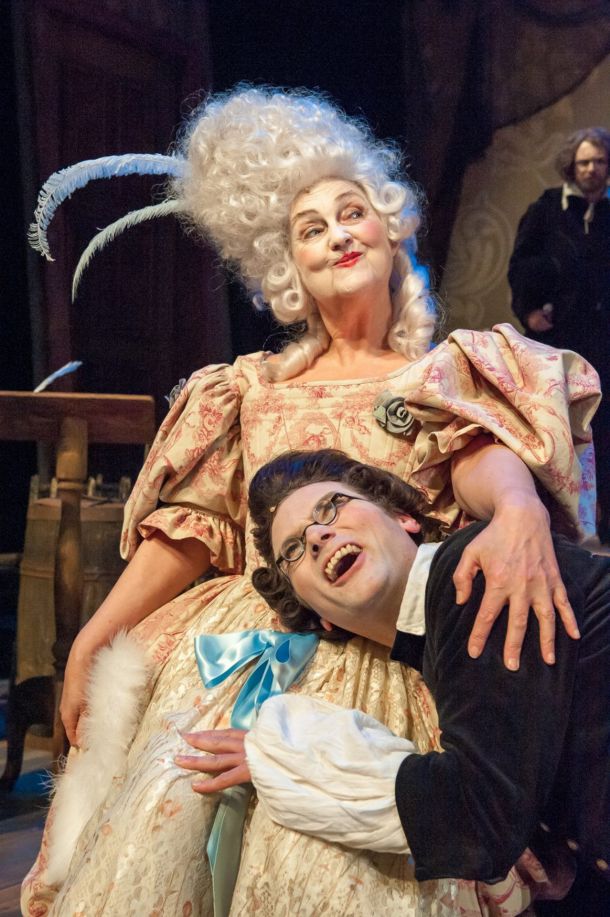 En mann og en kvinne i barokke kostymer, parykker og sminke. Mannen har hodet sitt mot kvinnens mage.