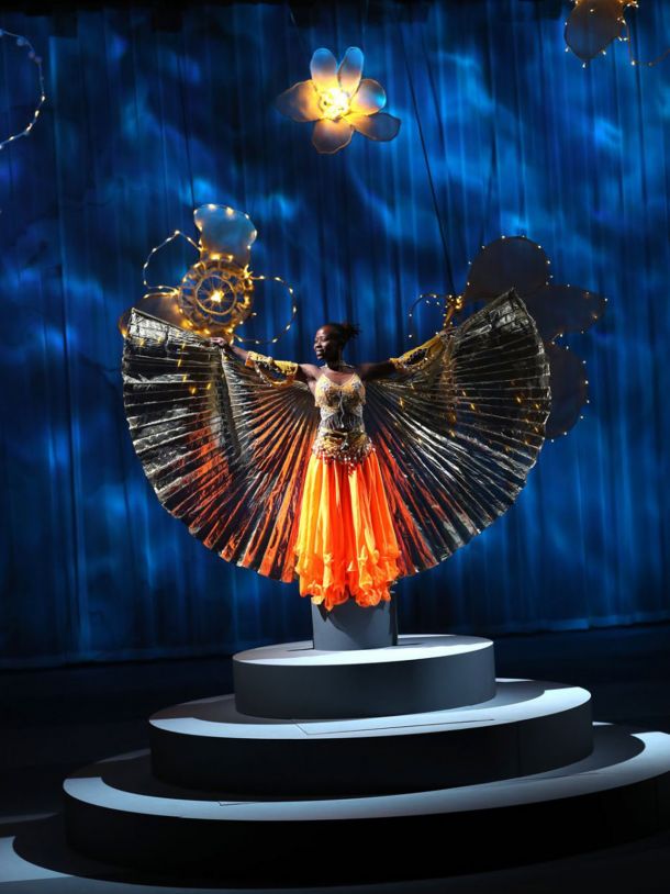 En ung mørkhudet kvinne står på en pidestall og strekker armene ut. Hun har på seg en fragerik ornasje kjole der skjørtet blir også til vinger.