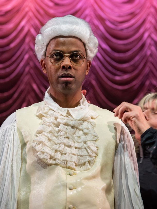 En mann kledd i hvite klær, skjorte og vest, med hvit parykk står å stirrer rett frem - et skarpt blikk. Han har briller på. En mann kledd som en dame står bak og retter opp på klærne.