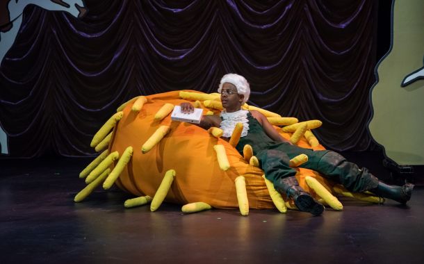 En mann med hvit parykk og skjeledress, sitter i det som skal være en høyhau.