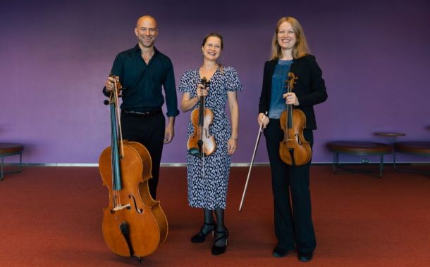 tre blide musikere som står på rødt teppe med lilla vegg bak seg, holder instrumentene i hånda og smiler