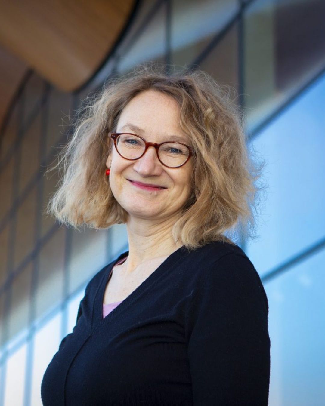 A woman with light hair and glasses smiling at the camera in front of a glass wall