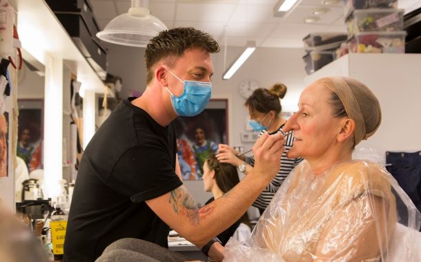 BIilde av mannlig sminkør som har på seg ansiktsmaske mens han sminker en kvinnelig skuespiller. Hun har fått påsatt lang nese og lang hake.