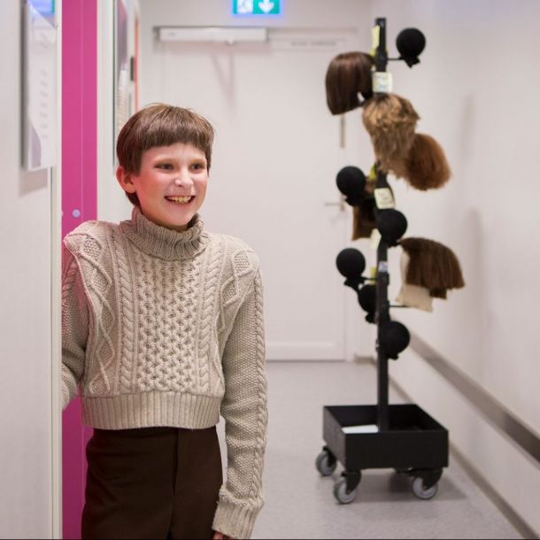 BIlde av gutt med pannelugg som smiler. Han står i en gang. I bakgrunnen skimtes parykker