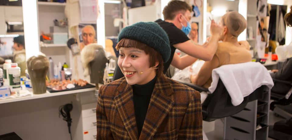 Bilde av ung kvinnelig skuespiller inne i en garderobe. I bakgrunnen skimtes en mann med munnbind som sminker en annen skuespiller.