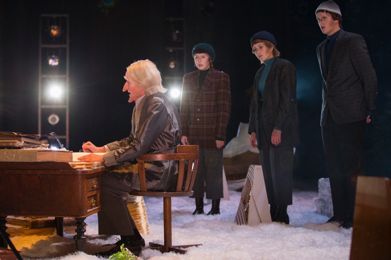 Bilde av gammel mann som sitter bak en skrivepult på en scene. I bakgrunnen står tre skuespillere i kostyme.