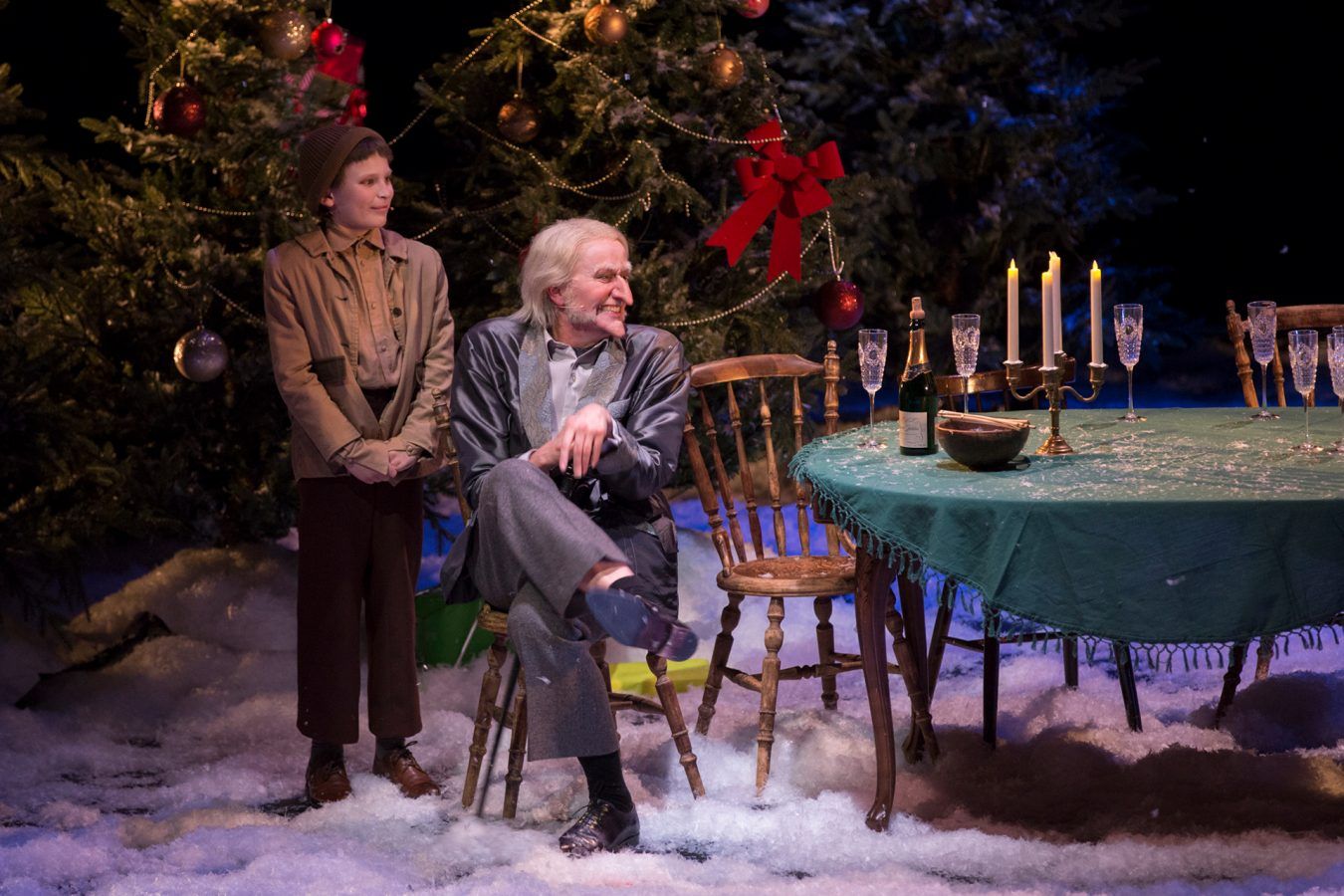 To skuespillere på en scene. Liten gutt står bak en gammel mann som sitter ved et bord.