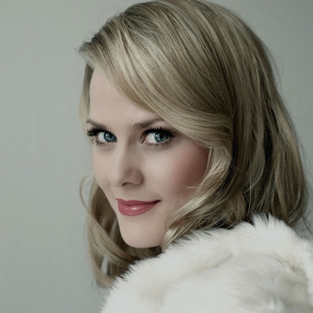 Image of a woman with long blonde hair looking into the camera and smiling. She is wearing a white fur coat