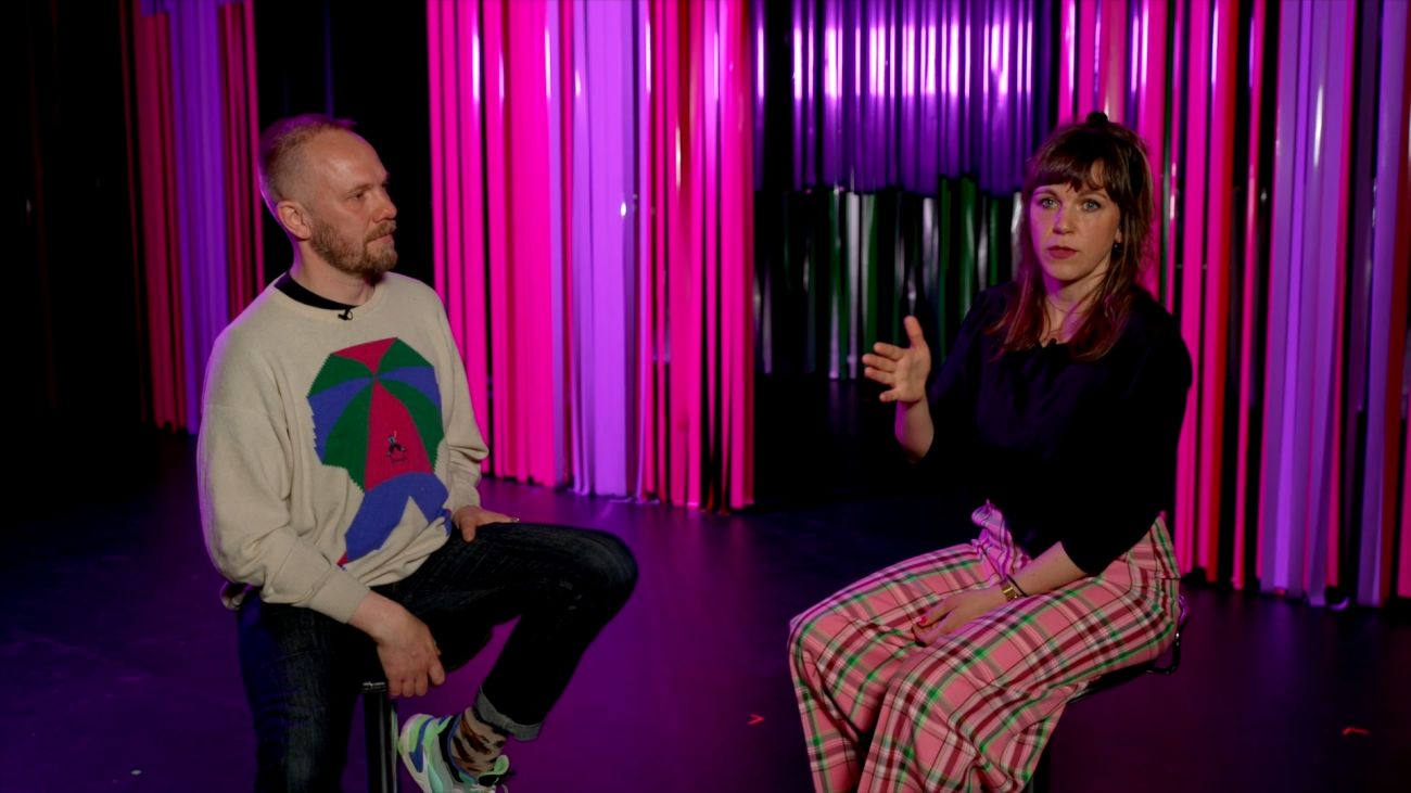 Bilde av mann og kvinne som sitter på to barklrakker. I bakgrunnen er det rosa og lilla remser som henger ned.