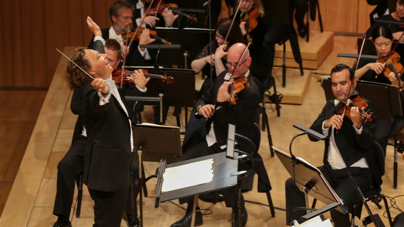 Bilde av kvinnelig dirigent som smiler mens hun strekker armene ut. Musikere som spiller i bakgrunnen.