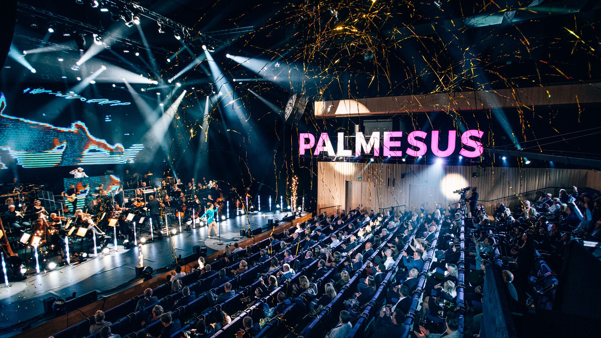 Bilde av publikum og scene med artister. Logo for Palmesus synlig i rosa i bildet.