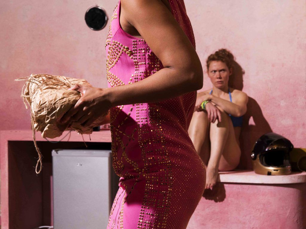 I forgrunnen ser du et menneske gå forbi i rosa kjole med en pakke i hendene. I bakgrunnen en kvinne i bikini som sitter på en benk med en scooter-hjelm ved siden av seg.